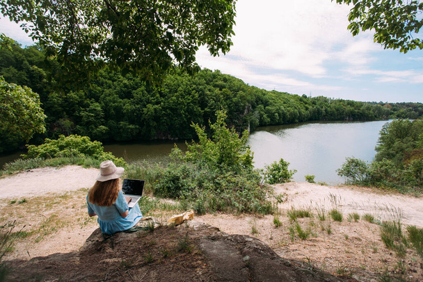 Die Freiberuflerin arbeitet mit einem Laptop in der Natur. Bekleidet mit einem blauen Kleid und einem hellen Hut. Sitzt auf Steinen in einem Waldgebiet, vor dem Hintergrund eines Flusses. In der Nähe liegt eine beige Ledertasche. - Foto, Bild