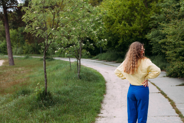 Une jeune fille aux cheveux bouclés blonds vêtus d'un jean bleu et d'une chemise jaune, portant des lunettes de soleil, se tient sur un chemin dans un parc pittoresque en été. - Photo, image