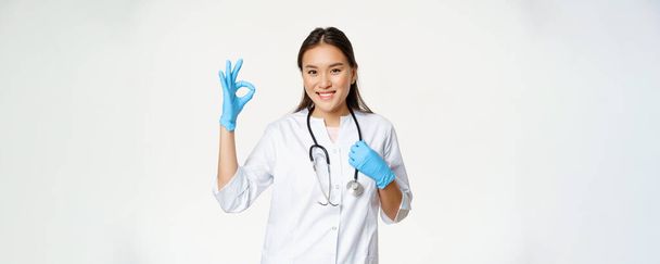 笑顔の医療従事者,ゴム手袋と医療制服のアジアの女性医師,承認を示しています,大丈夫なサイン,白い背景. - 写真・画像