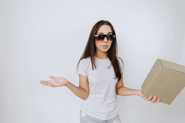 young woman holds cardboard box in her hands. Girl Standing Against White Background in Basic T-Shirt and Sunglasses. Small Business Concept, Place Under Text - Photo, Image