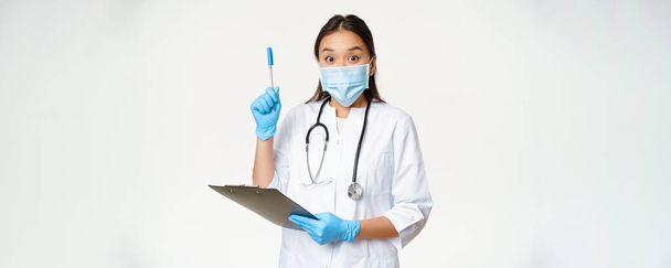 Ενθουσιασμένη γυναίκα γιατρός, Ασιάτης γιατρός κρατώντας πρόχειρο και την αύξηση στυλό, βρέθηκε λύση ή ιδέα, στέκεται σε ιατρική μάσκα προσώπου πάνω από λευκό φόντο. - Φωτογραφία, εικόνα