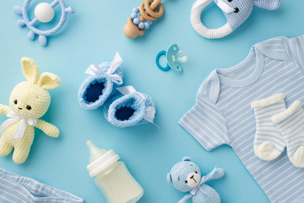 Baby accessories concept. Top view photo of infant clothes blue shirt knitted booties socks bunny and teddy-bear toys teether rattles milk bottle and pacifier on isolated pastel blue background - Photo, Image