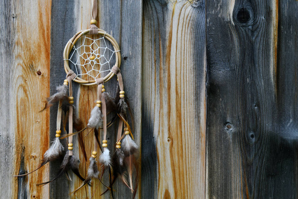 Nahaufnahme eines kleinen Traumfängers mit braunen Federn, der an einem alten verwitterten Holzzaun hängt.  - Foto, Bild