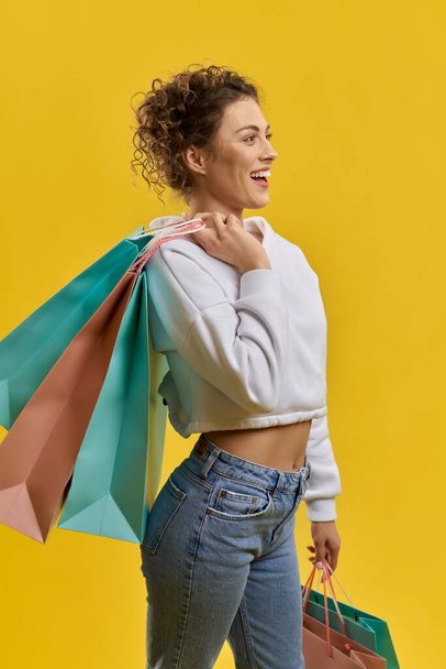 Confident curly woman carrying shopping bags on shoulder, smiling indoor. Side view of slim female shopper looking happy, isolated on orange studio background, with copy space. Concept of shopping.  - Foto, Bild