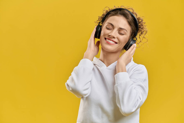 Delighted woman with black earmuffs listening music, having rest indoor. Portrait view of focused female enjoying sound, with closed eyes, smiling, isolated on orange background. Concept of enjoyment. - Φωτογραφία, εικόνα