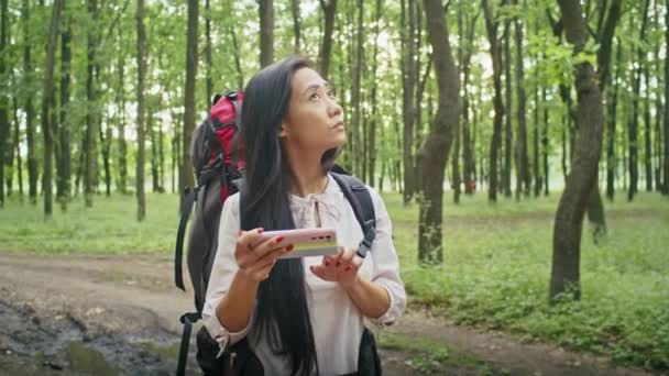 mujer asiática feliz con una mochila buscando una manera en el bosque, usando un teléfono, hablando en una videollamada, mirando un mapa. camping en verano - Imágenes, Vídeo