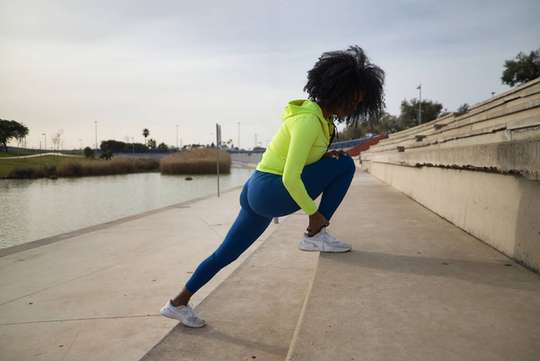 African-American woman with afro hair and sportswear, with fluorescent yellow sweatshirt and leggings, doing leg stretches on stairs. Fitness concept, sport, street, urban. - Фото, изображение