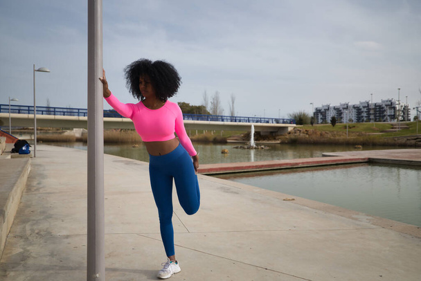 African-American woman with afro hair and sportswear, with fluorescent pink t-shirt and leggings, doing leg stretches holding on to a lamppost outdoors. Fitness concept, sport, street, urban. - Фото, изображение