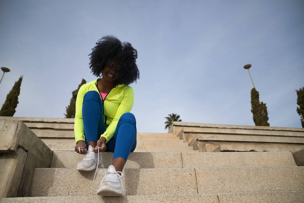 Afroameričanka s afro vlasy a sportovním oblečením, sedící, v kamaších, připínající tkaničky na cvičení na ulici. Fitness koncept, sport, ulice, městský. - Fotografie, Obrázek