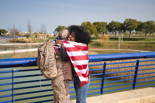 Афроамериканка с американским флагом обнимает своего партнера, американского солдата, только что вернувшегося с миссии. Армия, миссии, военные, война, любовь, объятия. - Фото, изображение