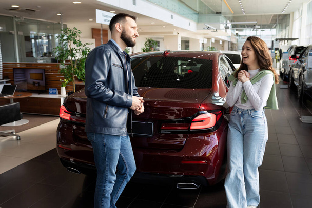 uma jovem mulher convence seu homem em um concessionário de carros para comprar-lhe um carro novo. - Foto, Imagem