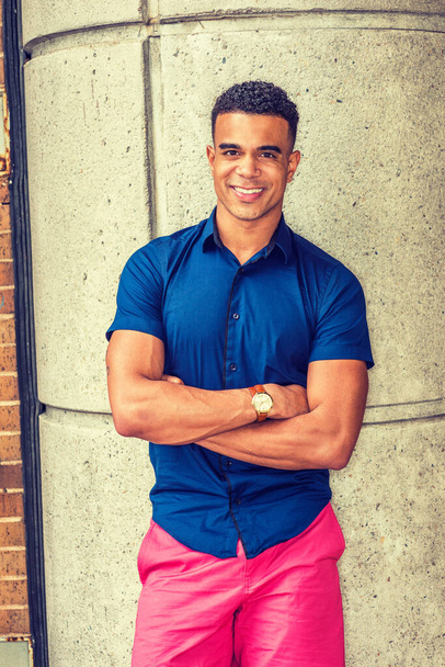 Portrait of Young African American Man in New York, wearing blue short sleeve shirt, red short, wristwatch, standing against concrete column on street, crossing arms, smiling.  - Foto, afbeelding