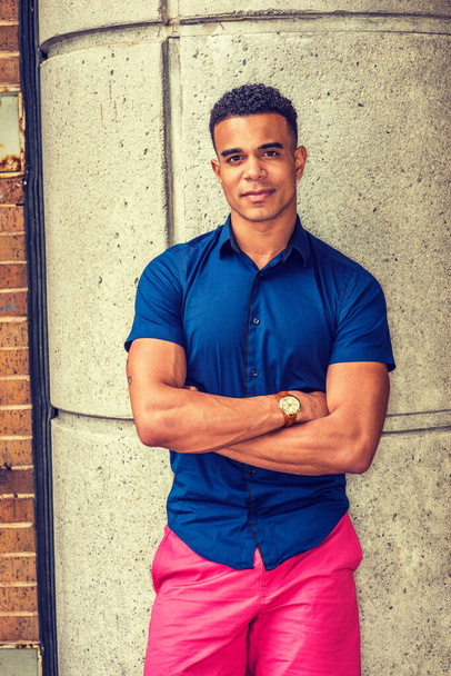 Nuoren afroamerikkalaisen miehen muotokuva New Yorkissa, yllään sininen lyhythihainen paita, punainen lyhyt, rannekello, seisoo betonipylvästä vasten kadulla, ylittää käsivarret, hymyilee.  - Valokuva, kuva