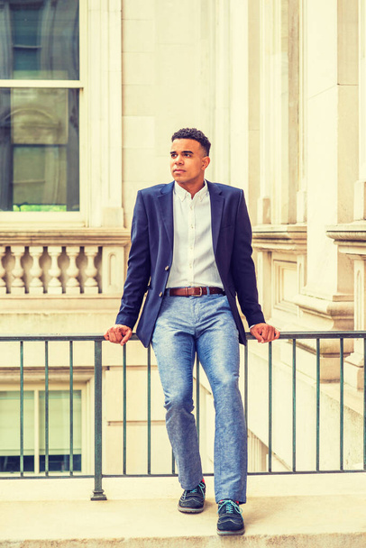 Het dragen van blauwe gloed, wit shirt, grijze broek, sneakers, Afro-Amerikaanse mannelijke student zit op leuning in vintage kantoorgebouw op de campus in New York, het nemen van pauze, denken.  - Foto, afbeelding