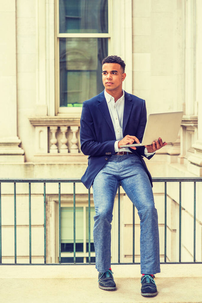 Afro-Amerikaanse student studeert in New York. Blauwe blazer, grijze broek, sneakers, kerel zit op leuning in vintage kantoorgebouw op de campus, werkt op laptop computer. Gefilterd effect. - Foto, afbeelding