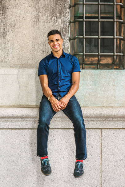 Retrato de Happy African American Man em Nova Iorque. Vestindo camisa de manga curta azul, jeans, meias vermelhas, tênis, relógio de pulso, cara se senta na parede vintage por janela, sorrisos.  - Foto, Imagem