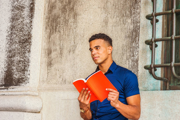 Uwielbiam czytać książki. Studia Afroamerykańskiego Studenta w Nowym Jorku. Młody człowiek w niebieskim krótkim rękawie, siedzi na kampusie pod zabytkową ścianą, czyta czerwoną książkę, patrzy w górę, poważnie myśli. - Zdjęcie, obraz