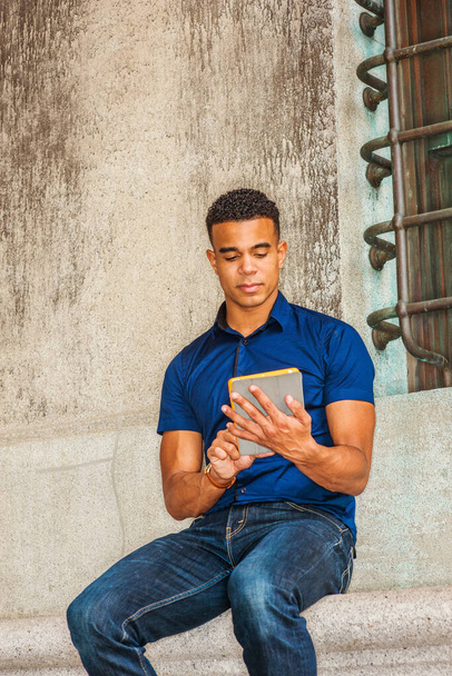 Я люблю читать. Современное чтение. Носить синюю рубашку с коротким рукавом, джинсы, афроамериканский студент колледжа, сидящий напротив старинной стены на улице в Нью-Йорке, читающий маленький планшетный компьютер. - Фото, изображение