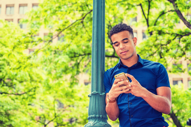 Afrikkalainen amerikkalainen opiskelija tekstailee ulkona, pukeutuu siniseen lyhythihaiseen paitaan, seisoo valopylvästä vasten kadulla New Yorkissa, lukee viestejä matkapuhelimella.  - Valokuva, kuva