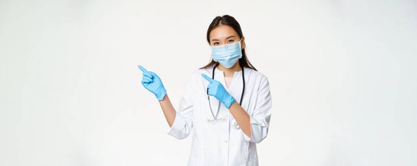 Retrato de una trabajadora médica asiática señalando con los dedos hacia la izquierda, usando mascarilla y guantes de goma, de pie en uniforme de la clínica sobre fondo blanco. - Foto, Imagen