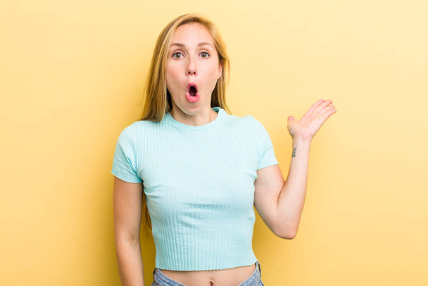 young adult  blonde woman looking surprised and shocked, with jaw dropped holding an object with an open hand on the side - Foto, imagen