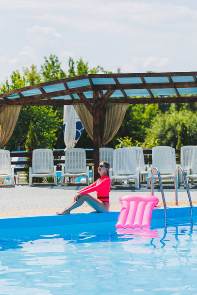 A beautiful woman in a pink swimsuit and with an inflatable mattress stands near a large pool. Summer holidays in a hotel with a swimming pool. Young girl in swimsuit and sunglasses - Valokuva, kuva