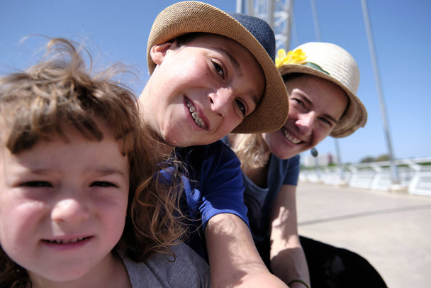 Adolescente feliz sonriendo y tomando selfie con mamá y hermano mientras pasa el fin de semana en el puente contra el cielo azul sin nubes en el día soleado - Foto, Imagen