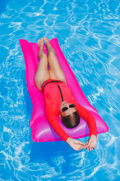 Mooi meisje in een roze badpak ontspannen op een opblaasbare roze matras in het zwembad. Een slanke hete vrouw in een zonnebril en badpak is aan het zonnebaden. Een vrouw ontspant zich in een luxe resort. - Foto, afbeelding