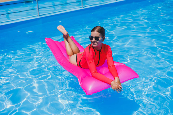 Attractive woman in sunglasses is relaxing and sunbathing on a pink mattress in the pool. Young woman in a pink swimsuit floats on an inflatable pink mattress - Photo, Image