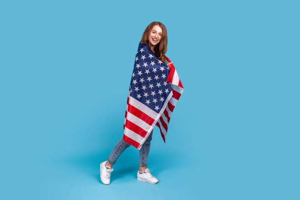 Full length portrait of smiling woman wearing striped sweater, standing wrapped in American flag, celebrating labor day, government employment support. Indoor studio shot isolated on blue background. - Fotografie, Obrázek