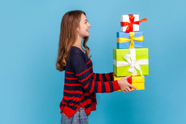 Side view portrait of smiling woman wearing striped sweater, holding out gift boxes, giving many present, congratulating with holidays, looking ahead. Indoor studio shot isolated on blue background. - 写真・画像
