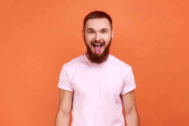 Portrait of bearded man showing out tongue and looking at camera with naughty disobedient grimace, making face, wearing pink T-shirt. Indoor studio shot isolated on orange background. - Φωτογραφία, εικόνα