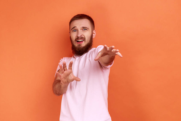 Portrait of bearded man raising hands from sudden fear, showing stop, no, refusal gesture, looking frightened, denying problems, wearing pink T-shirt. Indoor studio shot isolated on orange background. - Valokuva, kuva