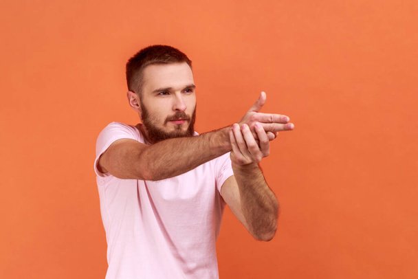 Side view of serious brave bearded man holding fingers pretending holding gun, aiming enemy, self-defense, wearing pink T-shirt. Indoor studio shot isolated on orange background. - Zdjęcie, obraz