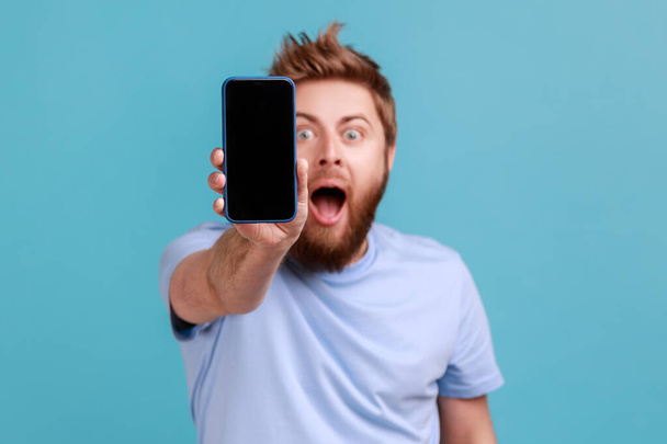 Portrait of bearded man holding out cellphone and looking at camera with big eyes and open mouth, saying wow surprised about technology. Indoor studio shot isolated on blue background. - Foto, Imagen