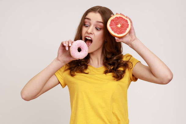 Hungry teenager girl in yellow T-shirt biting sweet junk donut instead of fresh juicy orange, choosing junk food, lack of willpower and motivation. Indoor studio shot isolated on gray background. - Foto, imagen