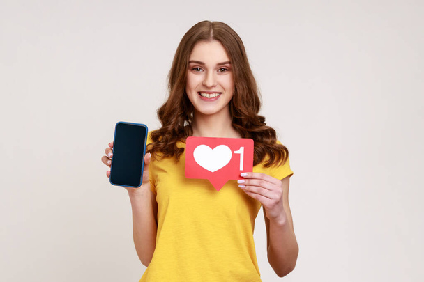 Positive smiling teen girl blogger holding like icon, showing smartphone with blank screen making post in social network and waiting for feedback. Indoor studio shot isolated on gray background. - Foto, imagen