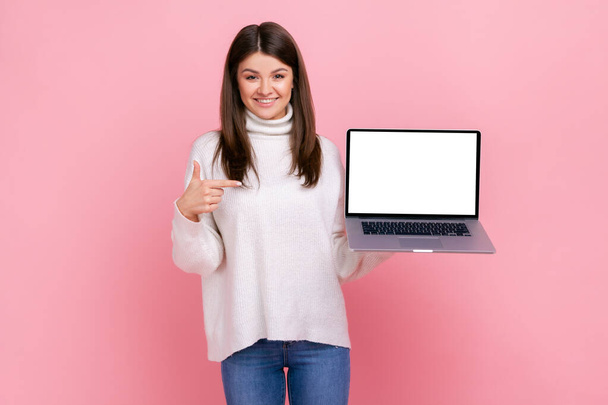 Gut aussehende Frau mit dunklen Haaren hält Laptop in den Händen, zeigt mit dem Finger auf leeres Anzeigendisplay, trägt weißen lässigen Pullover. Indoor-Studio isoliert auf rosa Hintergrund aufgenommen. - Foto, Bild