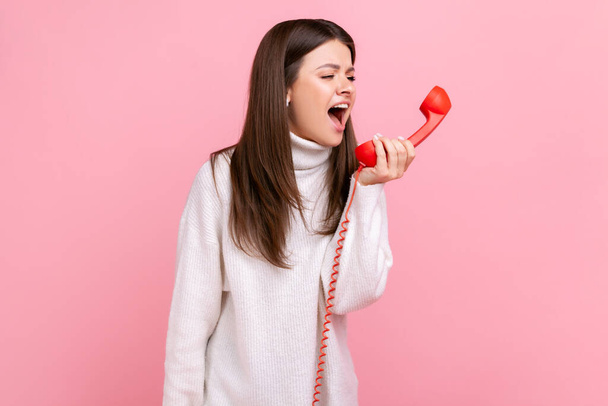 Angry young brunette woman screaming on red retro handset, expressing aggression, annoying client, wearing white casual style sweater. Indoor studio shot isolated on pink background. - Foto, afbeelding