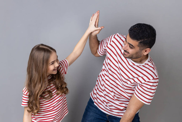 Portrait of playful cheerful father and daughter in striped T-shirts standing together, little girl giving high five to happy dad. Indoor studio shot isolated on gray background. - Foto, afbeelding