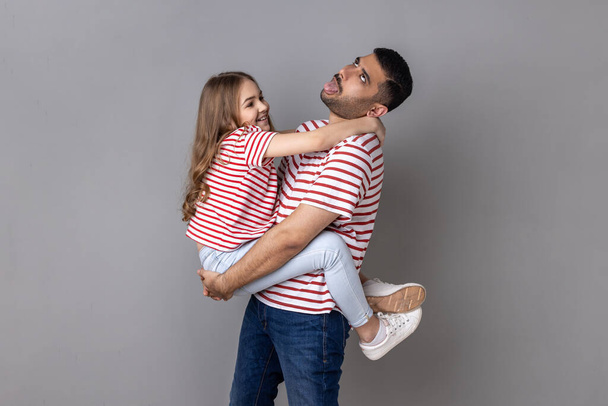 Portrait of funny father and smiling charming daughter in striped T-shirts standing together, tired dad holding kid and showing tongue out. Indoor studio shot isolated on gray background. - 写真・画像