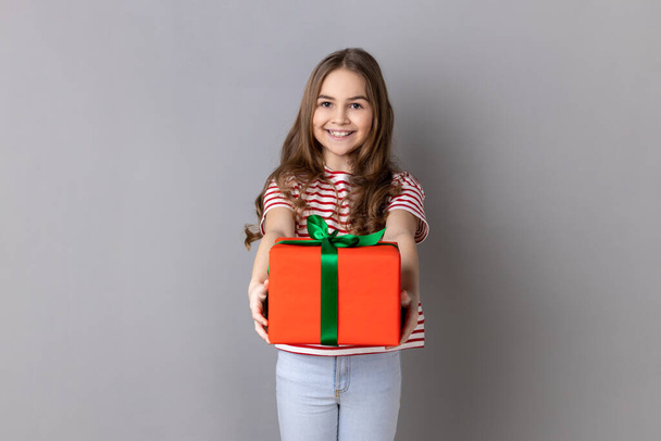 Portrait of smiling little girl wearing striped T-shirt giving present and congratulating on birthday, mother's day, offering gift box for charity. Indoor studio shot isolated on gray background. - Φωτογραφία, εικόνα