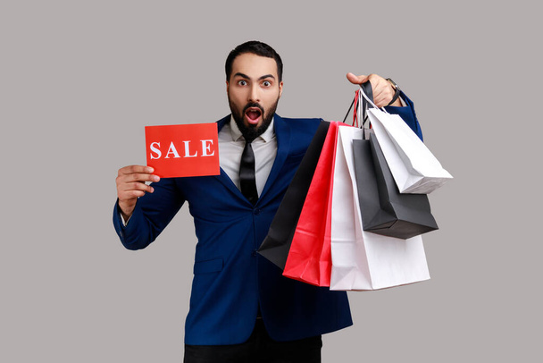 Bearded man astonished boasting with purchases holding paper shopping bags and sale card, big holidays discounts, wearing official style suit. Indoor studio shot isolated on gray background. - Photo, Image