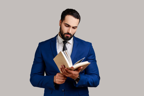 Concentrated bearded man reading book, enjoying reading, standing absorbed with exciting plot, checking schedule in notepad, wearing official style suit. Indoor studio shot isolated on gray background - Photo, Image
