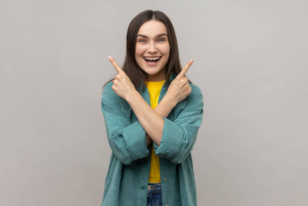 Portrait of positive woman with dark hair standing with crossed hands and pointing to copy space on both sides, wearing casual style jacket. Indoor studio shot isolated on gray background. - Foto, immagini