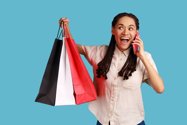 Portrait of excited amazed woman with black dreadlocks holding shopping bags and talking phone, boasting purchase to her friend, wearing white shirt. Indoor studio shot isolated on blue background. - Photo, Image