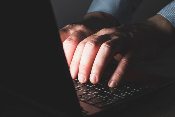 Le mani di un uomo caucasico irriconoscibile con una camicia blu che digita su una tastiera di un computer portatile. La scena è buia e le mani sono illuminate dalla luce dello schermo. - Foto, immagini