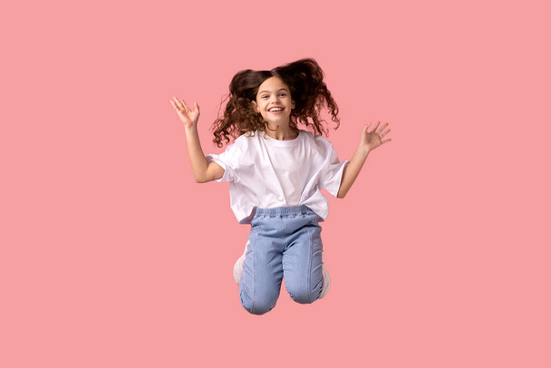Portrait of happy delighted positive little girl wearing white T-shirt jumping in air with raised arms, feeling energetic and lively. Indoor studio shot isolated on pink background. - Foto, imagen