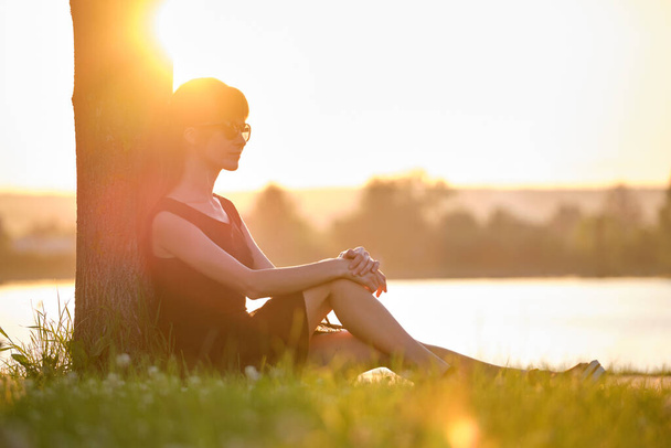 Mujer joven en traje casual sentado en césped de hierba verde que se relaja en el lado del lago en la noche caliente. Vacaciones de verano y concepto de viaje. - Foto, imagen