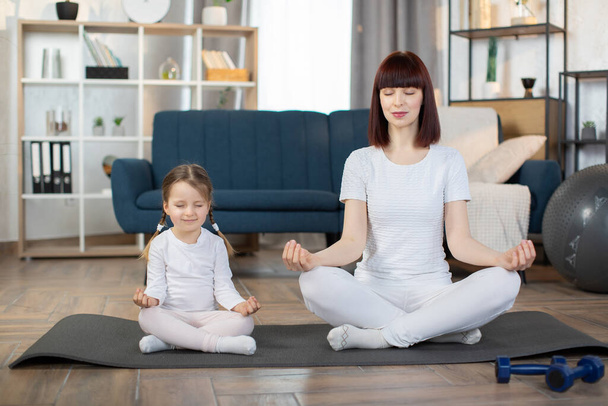 Beautiful young woman and her charming little daughter are smiling while doing yoga together at home. Mother and cute little girl smiling while sitting with closed eyes in lotus pose with on yoga mat - Foto, Imagem